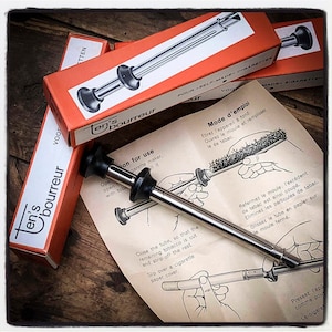 May include: A close-up of a silver cigarette roller with a black handle. The roller is on a white paper with instructions on how to use it. The instructions are in French and English. The text on the paper reads "Ten's Bourreur" and "Mode d'emploi".