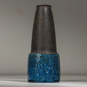 May include: A tall ceramic vase. The upper section is dark brown and speckled, while the lower section is textured turquoise blue. The vase has a cylindrical base and a conical top. The vase is a decorative item, suitable for displaying flowers or as a standalone piece.