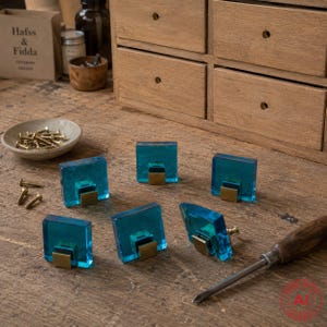 Peut inclure: Un ensemble de six boutons de placard carrés avec des faces en résine bleu sarcelle translucide et des bases en laiton brossé. Chaque bouton présente une face plate et un point de fixation central. Le lot inclut des vis métalliques pour la pose.