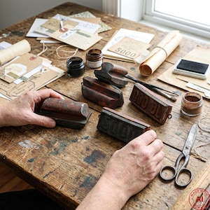 May include: A wooden table with vintage stamps, ink bottles, and stationery. A person's hands are holding a stamp, with other stamps, scissors, and rolled paper nearby. The items suggest a crafting or journaling theme, with a focus on aged materials.