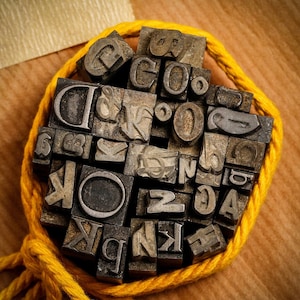 Könnte beinhalten: Eine Sammlung von Vintage-Metall-Letterpress-Druckblöcken in verschiedenen Größen und Stilen, die mit gelbem Garn zusammengebunden sind. Die Blöcke zeigen Buchstaben, Zahlen und Symbole, darunter die Buchstaben 'D', 'O', 'K', 'N' und 'A'.