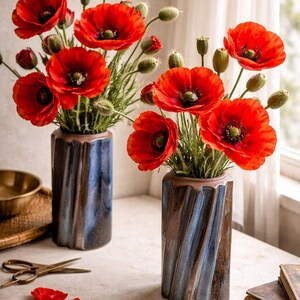Op de afbeelding: Twee keramische vazen met een blauw en bruin wervelontwerp bevatten levendige rode klaprozen. De bloemen hebben donkere centra en groene stelen. De scène omvat een schaar, bloemblaadjes en boeken.