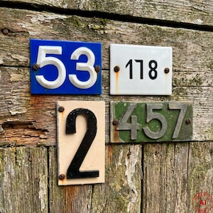 Op de afbeelding: Vier rechthoekige huisnummerplaten zijn gemonteerd op verweerd hout. De platen zijn blauw, wit, crème en groen en tonen elk een ander nummer in vetgedrukte, contrasterende kleuren. De nummers zijn 53, 178, 2 en 457.