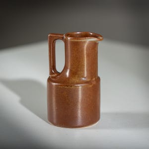 May include: A small, brown ceramic jug with a handle and a spout. The jug has a cylindrical base and a slightly flared top. The handle is rectangular and attached to the side. The jug is approximately 13 cm tall.