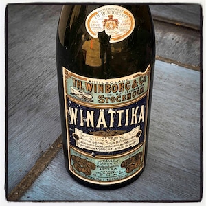 Può includere: Una bottiglia vintage in vetro scuro con un'etichetta dettagliata con il testo "WINÄTTIKA" e "TH. WINBORG & Co STOCKHOLM". L'etichetta ha uno schema di colori blu e oro con vari elementi di testo e illustrazioni.