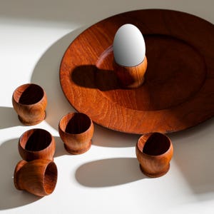 Puede incluir: Un juego de seis hueveras de madera con una bandeja de servir de madera. Las hueveras son marrones y tienen un acabado liso. La bandeja también es marrón y tiene una veta de madera natural. Un huevo está sentado en una de las hueveras.