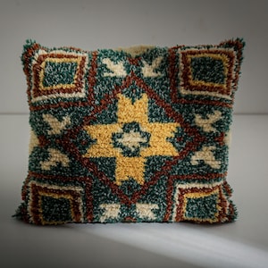 May include: Square decorative pillow with a textured, tufted design. The pillow features a central yellow starburst pattern, surrounded by teal, cream, and brown geometric shapes. The pillow is a square shape.