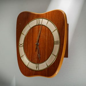 Könnte beinhalten: Eine Vintage-Wanduhr mit Holzrahmen und cremefarbenem Zifferblatt. Die Uhr hat römische Ziffern und goldfarbene Zeiger. Der Holzrahmen hat eine markante, geschwungene Form, typisch für das Mid-Century-Design.