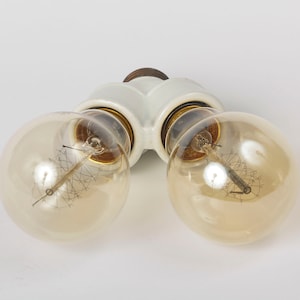 May include: A white ceramic double light fixture with two clear glass light bulbs. The bulbs have a filament design and are turned on.