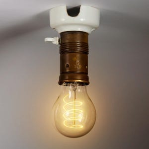 Peut inclure: Une ampoule vintage avec un filament en spirale, suspendue à un luminaire de plafond en porcelaine blanche avec une base en laiton. La base est marquée du mot "DAF".