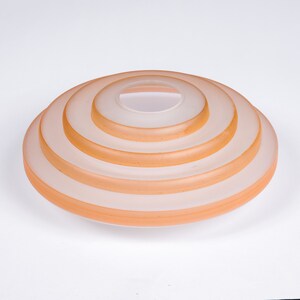May include: A set of five concentric glass rings, each with a slightly different shade of peach, creating a layered effect. The rings are stacked on top of each other, forming a circular shape.
