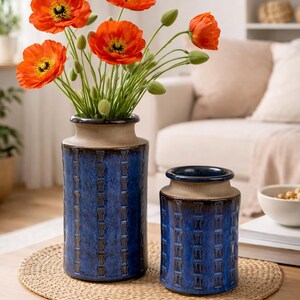 Könnte beinhalten: Zwei blaue Keramikvasen mit strukturiertem Muster und beigen Rändern, die leuchtend orange Mohnblumen enthalten. Die Vasen stehen auf einer gewebten Matte auf einem Holztisch und bilden einen dekorativen Blumenschmuck.