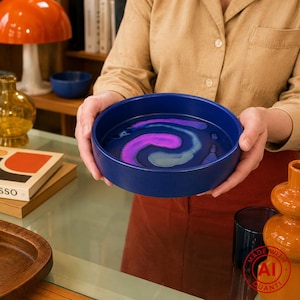 Op de afbeelding: Een ronde kom met diepe zijkanten en een matte marineblauwe buitenkant. De binnenkant heeft een glanzend, wervelend abstract patroon in tinten felpaars, lichtblauw en donkerblauw, wat een vloeiend marmerachtig effect op de bodem creëert.