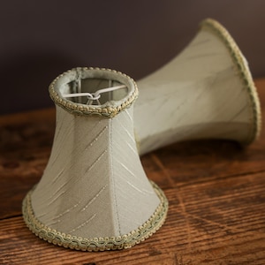 May include: Two sage green lampshades with a gold trim. The lampshades are made of a textured fabric and have a bell shape.