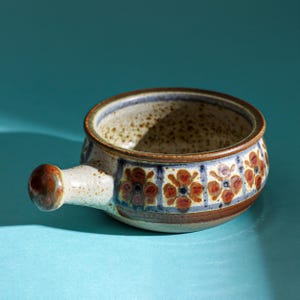 Op de afbeelding: Een klein, bruin keramiek serveerschaaltje met een handvat. Het schaaltje heeft een bloemmotief in tinten bruin, blauw en wit.