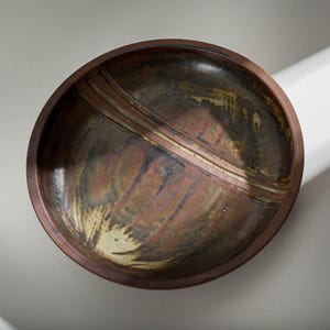 Può includere: Una ciotola in ceramica fatta a mano con un bordo marrone scuro e un design centrale con striature rosse, marroni e crema. L'interno ha una finitura lucida. I toni della terra della ciotola la rendono adatta per servire o esporre.