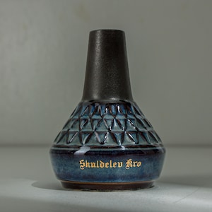 May include: A ceramic vase with a dark gray top and a blue patterned body. The body features a geometric design and the words "Skuldelev Kro" in gold lettering. The vase has a bulbous shape and a dark brown base.