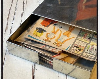 May include: A silver metal file box with a stack of vintage comic books and paper dolls inside. The comic books have black and white illustrations and the paper dolls are dressed in colorful outfits.