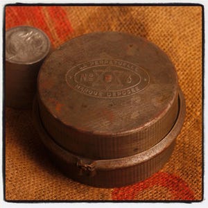 May include: A vintage, round, metal container with a hinged lid and a smaller cylindrical metal object to the left. The container has the text "LA PERPETUELLE" and "MARQUE DEPOSEE" with a star design. The objects are on a burlap surface.