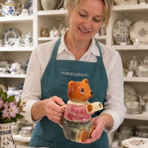 Könnte beinhalten: Eine braune und rosa Keramik-Teddybär-Teekanne mit weißem Ausguss. Die Person, die sie hält, trägt eine tealfarbene Schürze mit dem Text "PORCELAIN & CO." Im Hintergrund sind Regale mit Porzellangeschirr zu sehen.