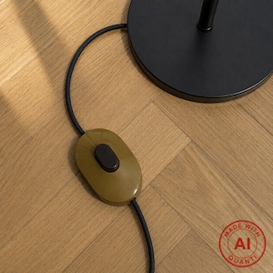 May include: A black circular lamp base connected to a black electrical cord featuring an olive green oval-shaped inline rocker switch. The switch has a small black button in the center for operation, and the cord extends from the base.