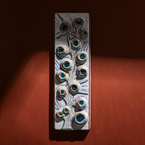 May include: A white ceramic wall hanging with a blue and white floral design. The design features a stylised plant with blue eyes.