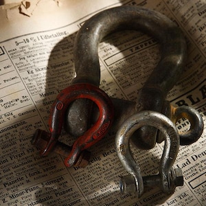 May include: Close-up of several metal shackles of varying sizes, with a weathered appearance. One shackle is painted red, and the others are silver. The items rest on a vintage newspaper.