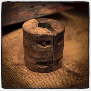 Könnte beinhalten: Ein verwittertes, zylindrisches Holzobjekt mit einem Spalt in der Mitte und einer gebogenen Kerbe oben. Das Holz hat ein dunkles, gealtertes Aussehen mit sichtbarer Maserung und kleinen Löchern. Ein Metallbolzen sichert die beiden Hälften.