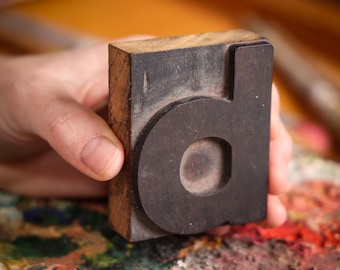 May include: A vintage letterpress printing block featuring the lowercase letter "d". The block is made of wood with a dark, circular letter form. The block is held in a hand, with a colorful paint palette in the background.
