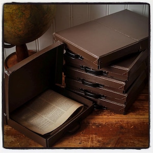 Peut inclure: Une pile de cinq boîtes de rangement rectangulaires en métal marron avec des poignées en plastique noir. Une boîte est ouverte, révélant une feuille de papier vieilli blanc cassé avec du texte imprimé. Les boîtes ont des coins renforcés.