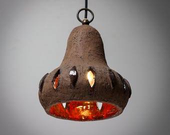May include: A brown ceramic pendant light with a bell-shaped shade and cut-out details. The light is illuminated, casting a warm glow through the cut-outs.