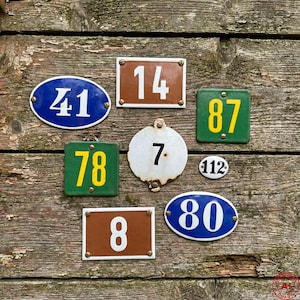May include: Collection of vintage metal house number signs in various shapes. Includes oval, rectangular, and circular signs with numbers in white or yellow against blue, green, brown, and white backgrounds. The signs are mounted on a weathered wooden surface.