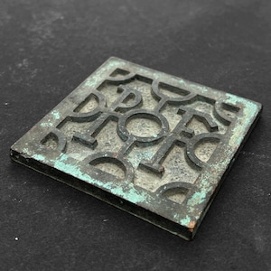 Puede incluir: Un azulejo cuadrado de metal con un patrón geométrico de círculos y líneas. El azulejo es de color verde oscuro con un acabado de pátina.