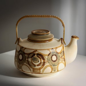 May include: Cream ceramic teapot with a brown and tan circular pattern. The teapot has a matching lid and a woven handle. The spout is curved, and the design is vintage.