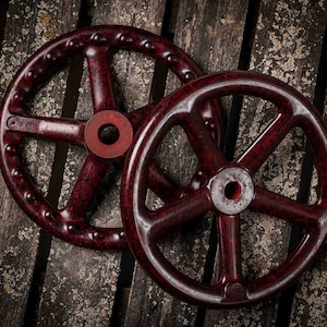Puede incluir: Dos ruedas de metal rojo oscuro con radios, una ligeramente más grande que la otra, están apiladas una encima de la otra. Las ruedas tienen un aspecto desgastado y envejecido y están colocadas sobre un fondo de madera rústico.