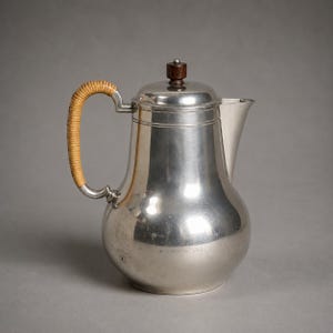 Könnte beinhalten: Ein silberfarbener Metallkrug mit bauchigem Körper, verjüngtem Hals und ausgestelltem Ausguss. Der Griff ist mit hellbraunem Flechtmaterial umwickelt, und der Deckel hat einen kleinen, dunkelbraunen zylindrischen Knopf in der Mitte.