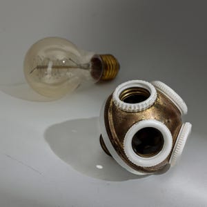 May include: A vintage-style light bulb and a multi-socket lamp holder. The bulb has a clear glass with a visible filament and a brass base. The lamp holder is white and gold with three sockets and a white trim.