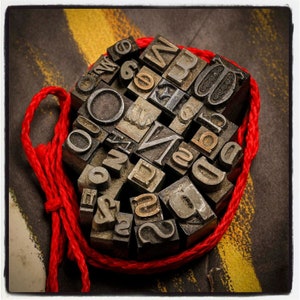 May include: A close-up of a collection of vintage metal letterpress printing blocks, tied together with a red cord. The blocks are arranged in a random pattern and feature a variety of letters and symbols.