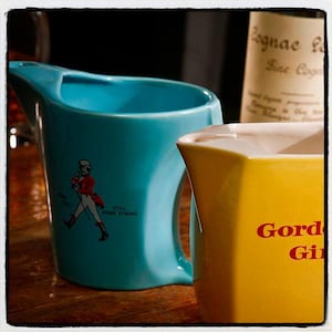 May include: Two ceramic jugs. One is turquoise with a handle and a graphic of a man in a red coat and the words "Still Going Strong." The other is yellow with the words "Gordon Gin" in red.