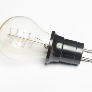 Op de afbeelding: Een vintage gloeilamp met een spiraalvormig filament en een zwarte bakelietstekker. De lamp is van helder glas en heeft een gele tint.