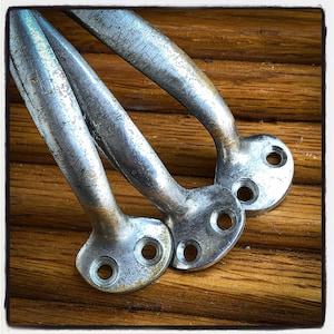 Può includere: Tre maniglie in metallo argentato e usurato, con un design curvo e due fori di montaggio ciascuna. Sono poste su uno sfondo di assi di legno marrone caldo, creando un'estetica rustica.