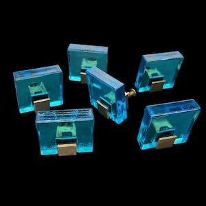 Op de afbeelding: Zes turquoise blauwe glazen kastknoppen met gouden metalen accenten. De knoppen zijn vierkant en rechthoekig.