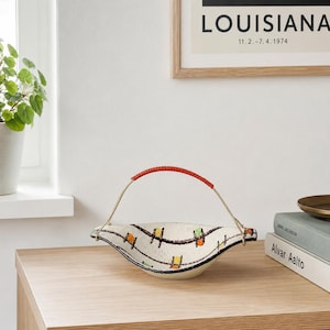 May include: A cream-colored ceramic basket with a red handle. The basket has a unique shape and is decorated with black lines and colorful square accents. The handle is made of a red material. The basket is sitting on a wooden surface.
