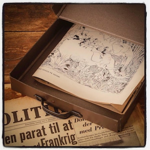 May include: A dark gray storage box with a handle containing a book with a black and white illustration of elephants and people in a jungle setting. The book cover features the text "NE SKULLE PÅ SAFARI" and rests atop a vintage newspaper.