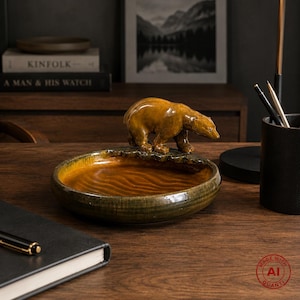 Pode incluir: Um prato redondo de cerâmica vidrada com um urso castanho esculpido na borda. O interior do prato tem uma textura ondulada semelhante à água em tons de âmbar e verde escuro, criando uma peça funcional para guardar pequenos objetos.
