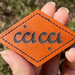 May include: A diamond-shaped leather patch with the word "ccca" embossed in black lettering.