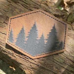 May include: A hexagonal leather patch with a brown background and a silhouette of five pine trees in black.