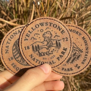 May include: Three brown leather patches with black stitching and text. The patches feature the names of national parks: Yellowstone National Park, Smoky Mountains National Park, and a third park with text that is partially obscured.