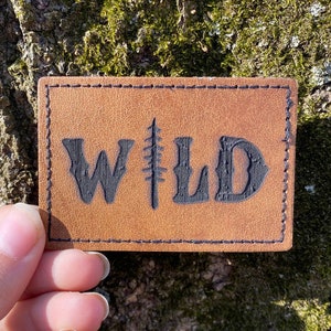 May include: A brown leather patch with black stitching and the word "WILD" in black lettering with a stylized tree design in the middle of the word.
