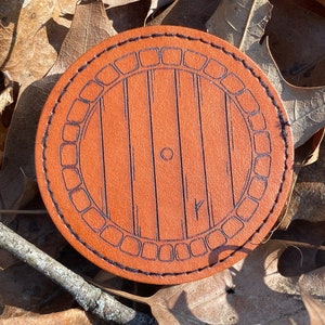 May include: A brown leather coaster with a black stitched border and a design of a round wooden door with a circular handle. The door has a wood grain pattern and is surrounded by a decorative border.